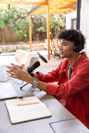 Man podcasting at patio table with mic laptop notebook pencil water glass headphones, copy space. Outdoor workspace, leisure, content creation, acoustic equipment, relaxed atmosphere, lifestyle, modern designの写真素材