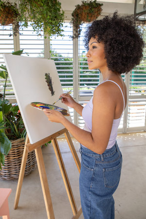 African American woman painting on easel at sunroom holding palette with paintbrush, copy space. Art, creativity, inspiration, botanical, craft, rustic, wellnessの写真素材