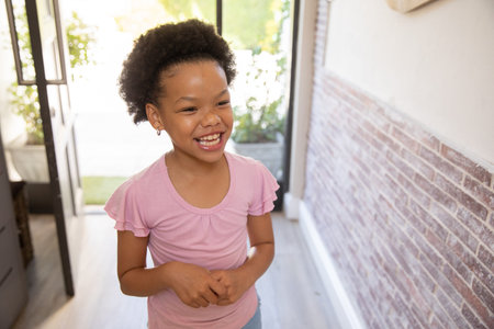 Smiling African American girl standing in entryway wearing pink shirt seeing garden via glass door. Children, interior design, minimalism, outdoor connection, natural light, domestic scene, lifestyleの写真素材