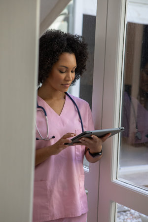 African American female nurse in pink scrubs with stethoscope standing at glass door reading tablet. Medical, healthcare, nursing, professional, clinical, technology, hospitalの写真素材