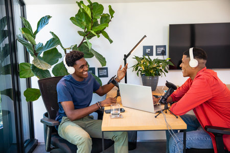 Diverse male co-hosts podcasting using microphones with laptop in modern podcast studio. Broadcast, communication, technology, innovation, teamwork, modern, creativeの写真素材