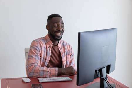African American man typing on wireless keyboard and smiling at computer monitor in home office. Professional, workspace, modern, technology, entrepreneur, focused, productivityの写真素材