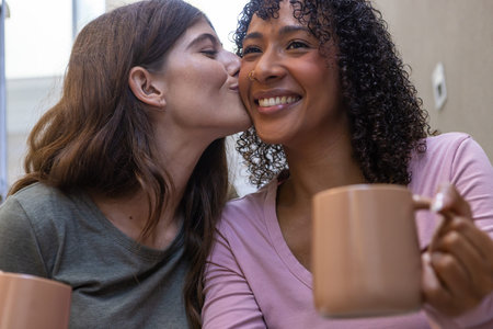 Diverse women sharing tender kiss while holding ceramic mugs at home near window. Romance, intimacy, friendship, warmth, connection, lifestyle, cozyの写真素材
