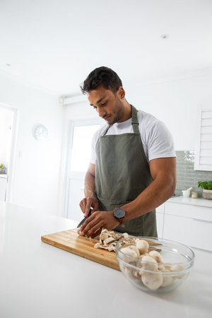 Male cook chopping mushrooms on cutting board with chef's knife at kitchen counter wearing apron. Home cooking, culinary, kitchenware, gastronomy, healthy living, rustic style, organicの写真素材