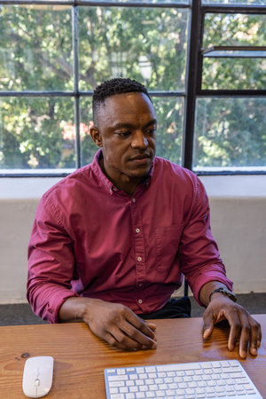 African American man sitting at wooden desk in office using wireless keyboard, mouse by grid window. Professional, workspace, modern, serene, contemplative, interior, greeneryの写真素材