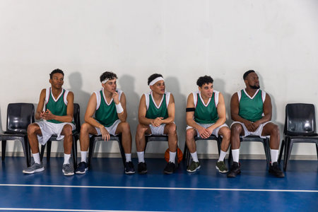 Diverse male basketball teammates waiting on chairs by court wearing jerseys with basketball nearby. Athletes, team spirit, sportswear, gymnasium, competition, camaraderie, dynamicの写真素材