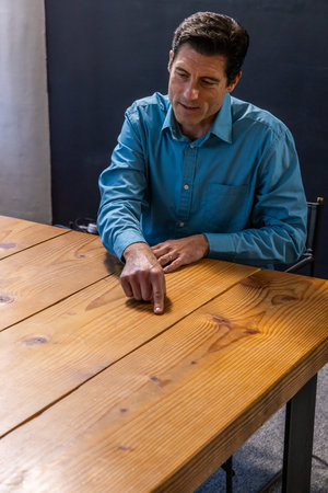 Middle-aged man wearing teal shirt pointing at wooden table grain in office, copy space. Professional, corporate, discussion, focus, workplace, seminar, collaborationの写真素材