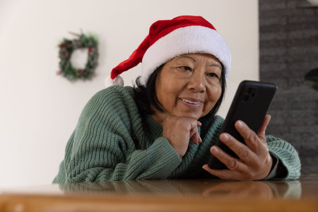 Senior Asian woman leaning on table at home checking smartphone near Christmas wreath and greenery. Festive, holiday, cozy, seasonal, modern, warm, lifestyleの写真素材
