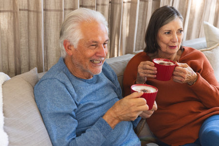 Senior couple sitting on beige sofa holding red ceramic mugs in living room with decorative pillows. Nostalgia, comfort, warmth, companionship, relaxation, cozy, domesticの写真素材