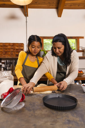 Diverse mother and daughter in aprons rolling dough with rolling pin on countertop in home kitchen. Family, collaboration, rustic, homely, warmth, skill, traditionの写真素材