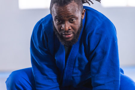African American man kneeling on blue padded mat inside martial arts studio wearing blue gi. Martial arts, athlete, training, discipline, focus, fitness, wellnessの写真素材