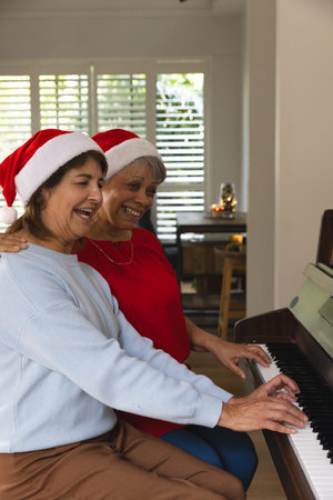 Senior female friends wearing Santa hats playing grand piano in living room with festive candle. Holiday, celebration, companionship, nostalgic, cozy, elegance, musicalの写真素材