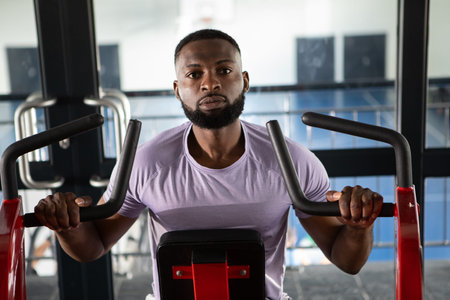 African American man pushing chest press handles at gym weight area wearing light purple tee....