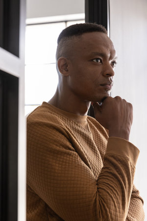 African American man resting chin, looking through frosted window in room wearing brown sweatshirt....
