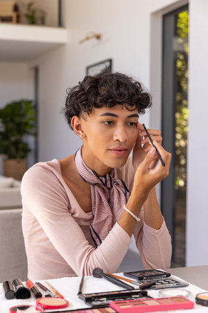 Man in his twenties applying eyebrow makeup with thin brush amidst eyeshadow palettes at home....