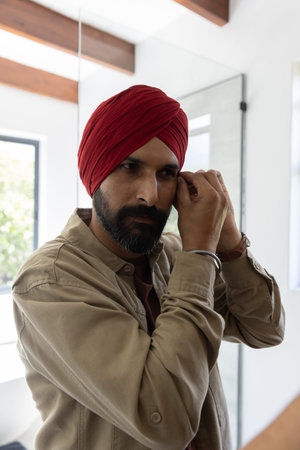 Asian Indian man adjusting earring at home bathroom mirror wearing red turban. Contemporary,...