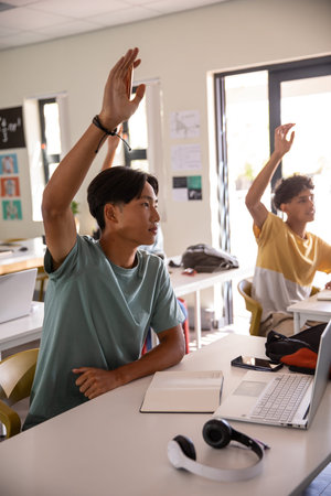 Teenage male classmates raising hands in classroom with laptops, notebooks, headphones, copy space....