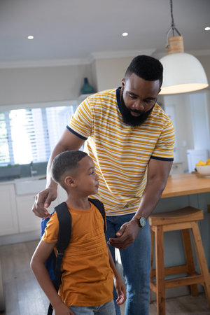 African American man and boy leaning over adjusting backpack straps at kitchen island with stools. Family, caregiving, home, domestic, nurturing, bonding, routineの写真素材