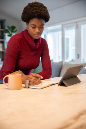 African American woman in red sweater writing in notebook referencing tablet at counter, copy space....