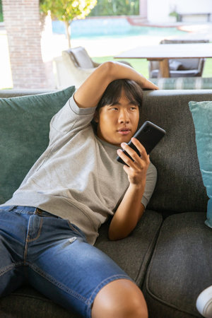 Asian teenage male reclining on padded sofa with teal pillows at backyard patio holding smartphone....