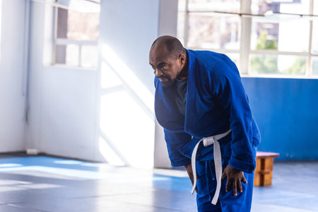 African American man bowing at dojo with blue gi, white belt near padded mats, copy space. Respect,...