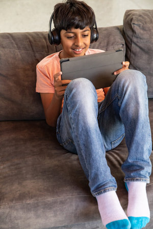 Indian boy sitting on grey sofa at home using tablet with headphones near cushions. Youth, leisure,...