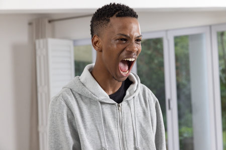 African American man shouting in living area by sliding glass doors, white shutter, pale wall. Energetic, expression, modern, casual, vibrant, emotion, relaxationの写真素材