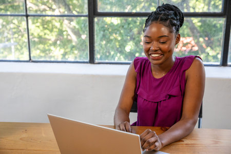 African American woman typing on laptop at desk in home office near window, copy space. Workspace,...
