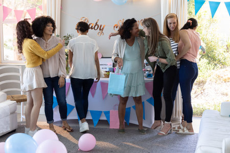 Diverse female friends celebrating baby shower in living room with pastel gift bags and balloons....