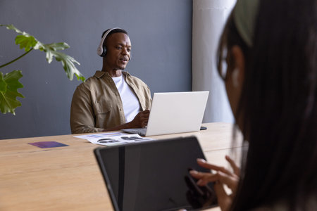 African American coworkers using laptop, tablet, headphones at wooden table with plant, copy space....