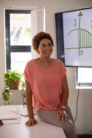 African American woman sitting on desk in modern office presenting graph beside laptop notebook....
