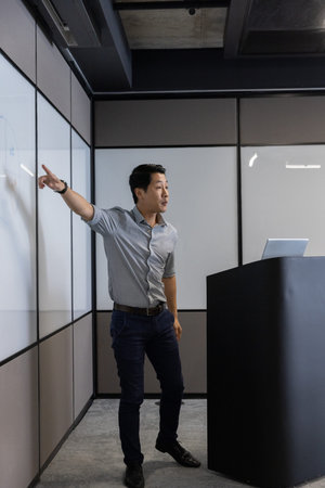 Presenter standing next to whiteboard and podium, using laptop while gesturing in meeting room....