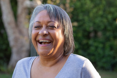 Senior African American woman laughing warmly while standing in backyard wearing light gray T-shirt....
