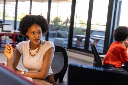 Diverse female coworkers sitting on ergonomic chairs at desk in office holding red pen by monitors. Professional, collaboration, workspace, modern, productivity, teamwork, corporateの写真素材