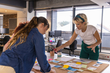 Diverse female coworkers examining sticky notes, mugs and printed diagrams on tabletop at office....