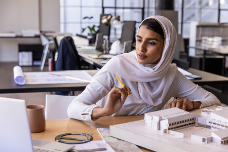 Asian Indian woman wearing hijab examining model at studio desk with laptop, pencil, copy space....