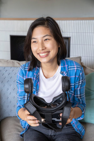 Asian woman sitting on grey couch in living room by fireplace holding virtual reality headset....