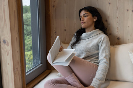 Hispanic woman sitting on bench seat with pillow reading paperback by window in wood-paneled nook....