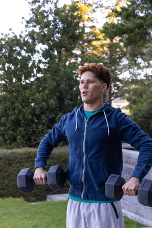 Male in blue hoodie and shorts performing dumbbell workout in backyard grass with evergreen trees....