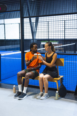 Diverse friends in sportswear resting on bench beside padel court holding coffee by padel racket. Athletes, leisure, athleticism, casual, mobility, teamwork, fitnessの写真素材