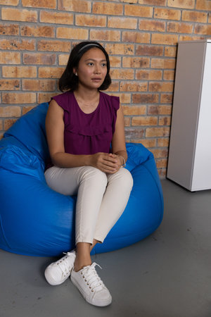 Asian woman sitting on blue bean bag chair by brick wall in lounge with white cabinet. Casual,...