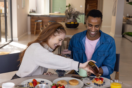 Diverse couple pouring syrup onto pancakes at wooden dining table at home with coffee press....