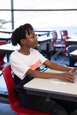 African American teen color-block tee studying on tablet at classroom desk by windows, copy space....