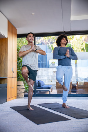 Diverse male friends practicing yoga balancing on one foot on black mats in modern living room. Wellness, fitness, mindfulness, harmony, relaxation, contemporary, lifestyle