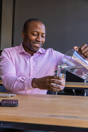 African American man pouring water into glass tumbler at table in office break room with smartphone....