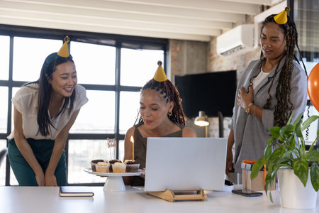 Diverse female coworkers celebrating birthday in office with laptop and cupcake stand with candle. Collaboration, celebration, modern, vibrant, teamwork, professional, cheerfulの写真素材