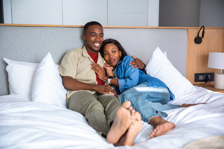 African American couple reclining together on bed in guest room with pillows and bedside lamp....