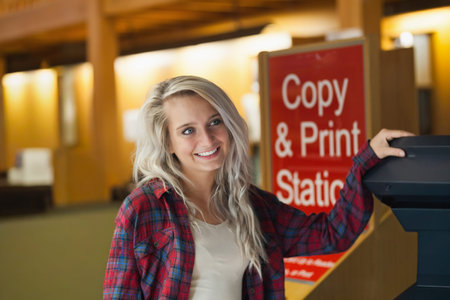 Female standing at library copy station near red sign and printer, wearing plaid shirt. Campus,...