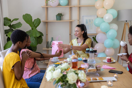 Diverse female friends handing pink gift bag to woman wearing sash at home under pastel balloons....