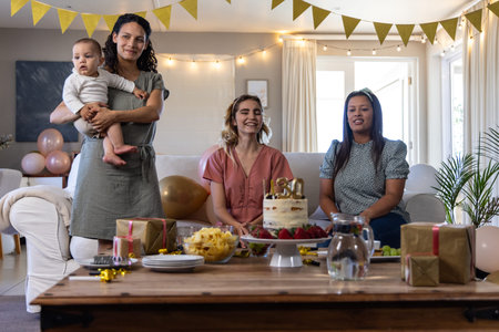 Diverse female friends gathering around table at home holding baby celebrating cake, balloons. Celebration, friendship, family, joy, domestic, pastel, cozyの写真素材